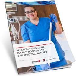 A hospital employee wearing scrubs and gloves and holding cleaning supplies.